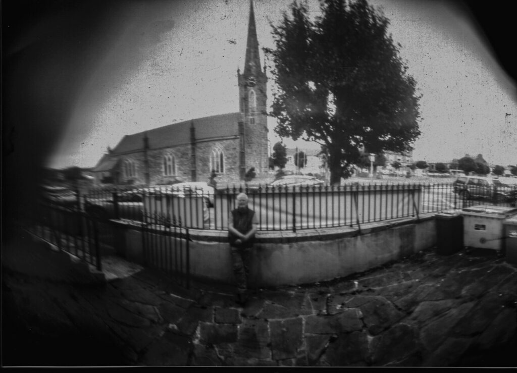 positive scan of black and white pinhole photograph (12 second exposure) showing a curator Ciarán Walsh standing against the wall of Kerry Writers' Museum with St John.s Theatre and Culture Centre - a repurposed church - in the background. The photo was taken through the aperture of a camera obscura using photographic paper wrapped around the inside of a cylindrical coffee tin hence the extreme curve or fisheye effect. Taken during a two-day workshop led by Cork-based artist Harry Moore as part of an analogue mini-fest Ciarán Walsh curated during Heritage Week 2025.