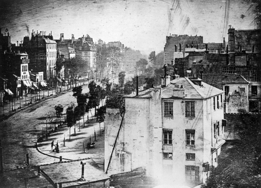 Louis Daguerre, 'Boulevard du Temple, Paris, 3rd arrondissement', Daguerreotype. Made in 1837 or 1838, this photograph is believed to be the earliest photograph that captured a living person. Shot from a roof, It captures a busy street in Paris, but because the exposure lasted for 4 to 5 minutes, anything moving traffic left no trace. Only the two men near the bottom left corner, one apparently having his boots polished by the other, stayed in one place long enough to be visible.