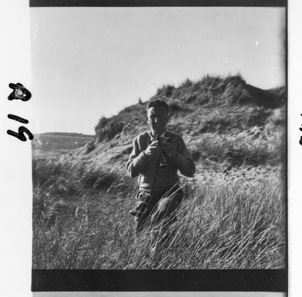 Digital scan Ciarán Walsh made in 2025 of a medium format black and white photograph shot around 1960. It shows a young man kneeling among sand dunes, with the sea just about visible in the background. This is Tony Fitzmaurice and he is holding a a Kodak Retinette camera and an earlier version of the same camera hangs from a strap around his neck. The photograph has a black border on three sides and this reveals that it is a detail of a riot of a sheet negatives, usually referred to as a contact sheet. Fitzmaurice wrote the number of each photograph in black marker. This is photo no. R19. It is one of 26,000 image in the Tony Fitzmaurice Collection. Kathy Reynolds gifted the collection to Kerry Writers' Museum in 20205 and Ciarán Walsh, Curator of Film and Digital Media at the museum, is currently digitising the collection (written October 2025).