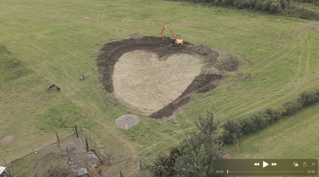 poster Image for Rushes | Luachair, an exhibition that celebrates women’s moving image art practice and features selected work by Lisa Fingleton, Laura Fitzgerald (project curator), Bláithín Mac Donnell, Lorraine Neeson, and Mieke Vanmechelen. The image was taken by drone and shows a large heart shape pond being excavated in a green field by a mechanical digger. Two donkeys look on. Lisa Fingleton took the shot. Ciarán Walsh posted it.
